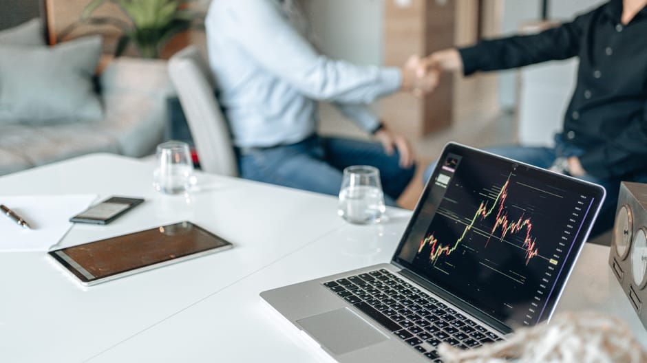 Two professionals shaking hands in an office with data analysis on a laptop screen.